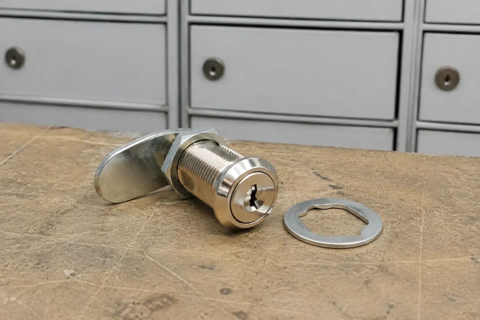 Detached threaded cam lock and mounting washer on a workbench in front of closed mail compartments. — Mailbox Lock Replacement, Waterloo