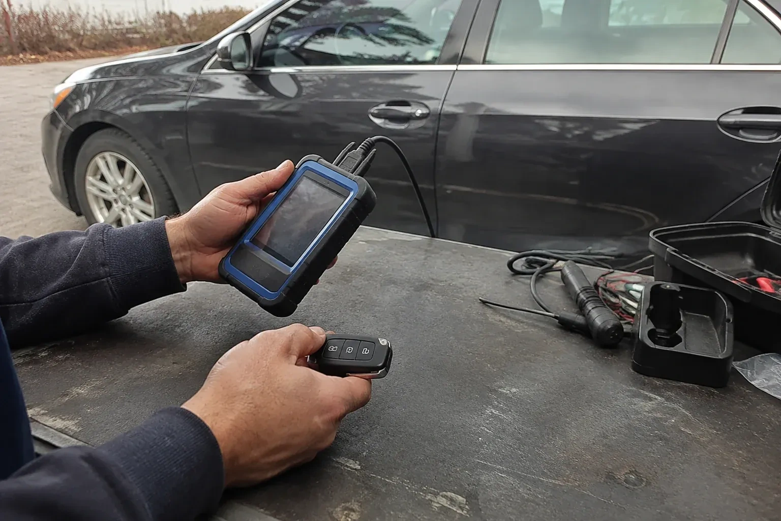 Hands holding a blue-edged electronic device and car-key fob at a worktable beside a parked car. — Transponder Key Programming, Waterloo