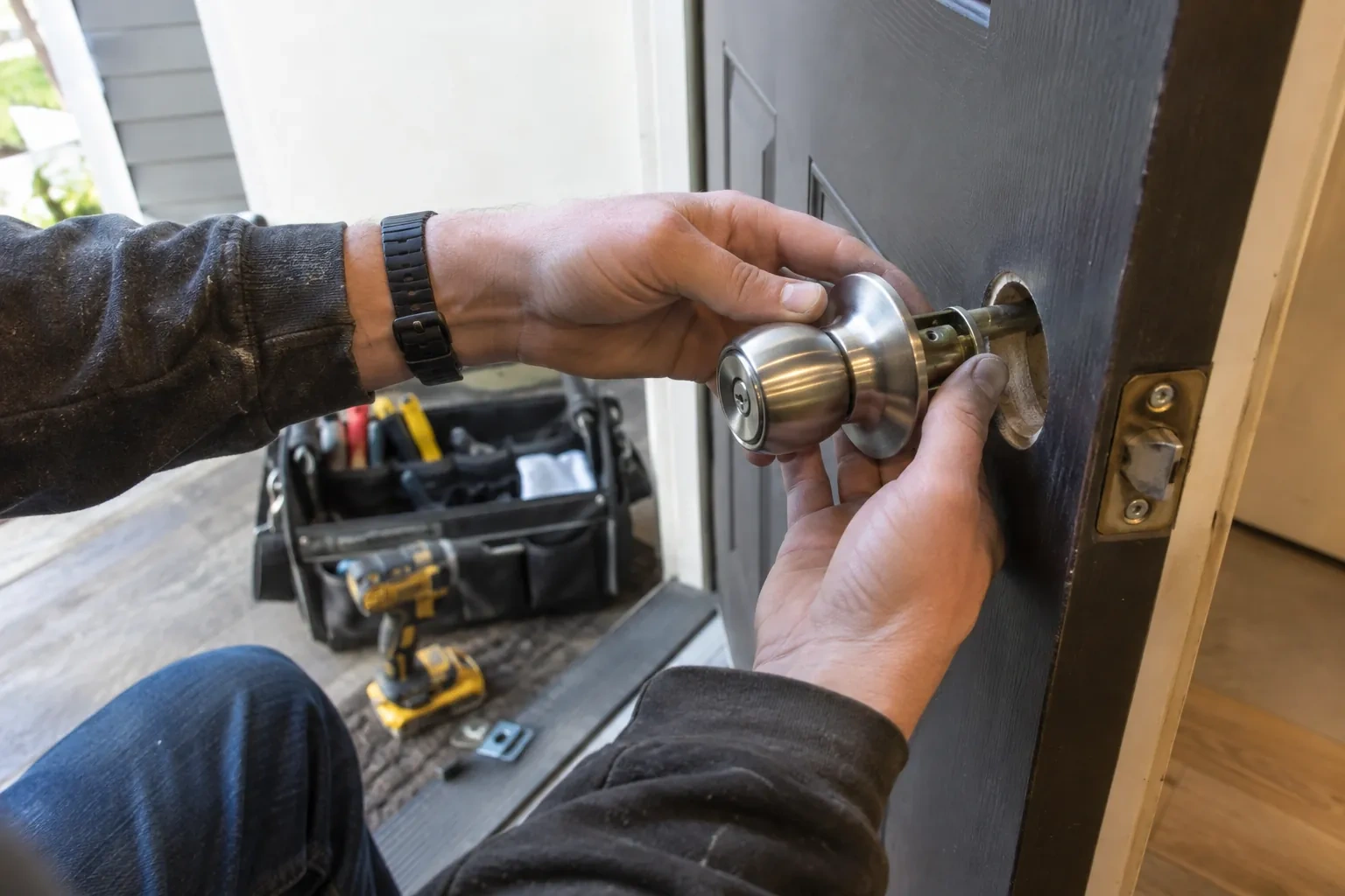 Hands fitting a steel door knob into the bore of a dark door, with a yellow drill and tool bag behind. — Hawkesville