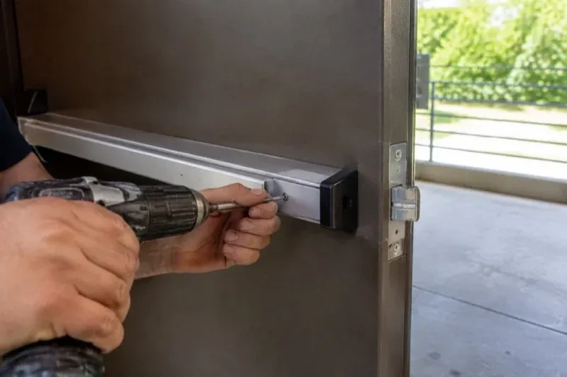 Hands using a power driver at the end of a horizontal push bar on an open metal door. — Panic & Exit Hardware, Waterloo